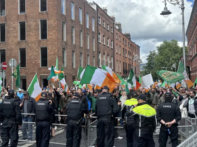 A large crowd of protesters gather near Leinster House in Dublin, carrying Irish tricolour flags. A line of Gardaí (Irish police) officers are facing the crowd, manning steel barriers.