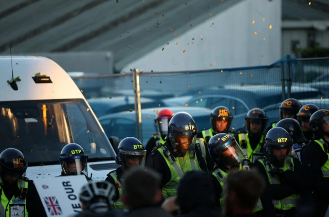 A line of officers wearing riot gear are standing in front of a riot van. Protesters are throwing large chunks of gravel which can be seen flying through the air
