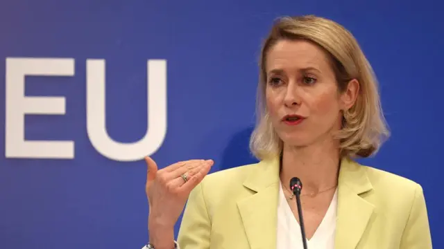 Kaja Kallas gestures as she speaks against a blue background with the letters 'EU' displayed. She is wearing a yellow jacket