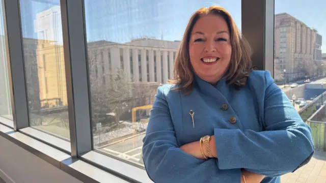 Tina McKenzie, a woman with shoulder-length brown hair, smiles at the camera with her arms folded. She is wearing a blue coat and is standing a large window overlooking a city centre street.