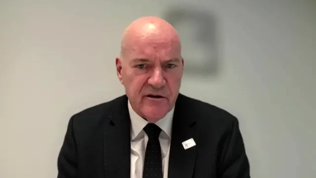 Kevin Craven sits in front of a white wall wearing a black suit