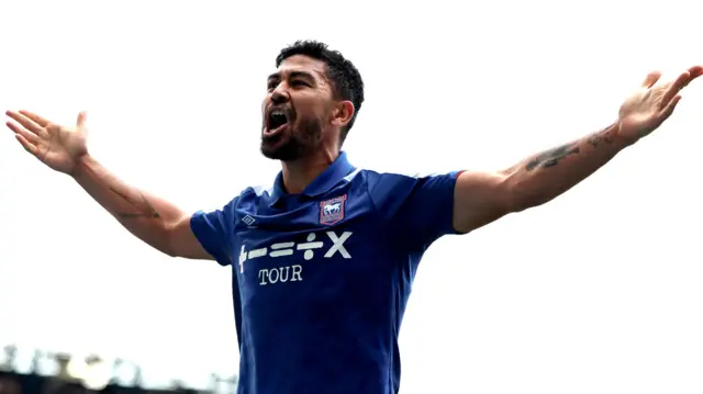 A football player celebrates scoring a goal. He wears a blue football shirt and has his arms raised out from his body as he shouts in celebration.