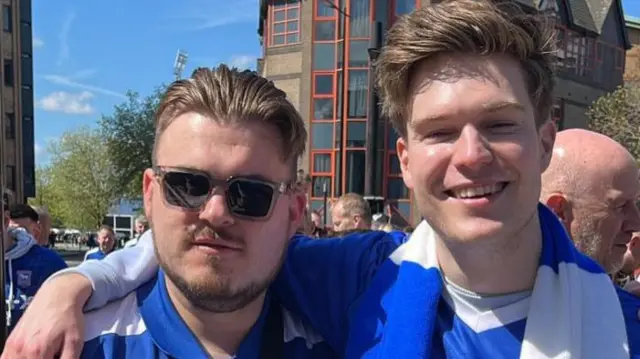 Two football fans stand together to pose for a photo. They wear matching blue football shirts. They both have short brown hair and one is wearing glasses.