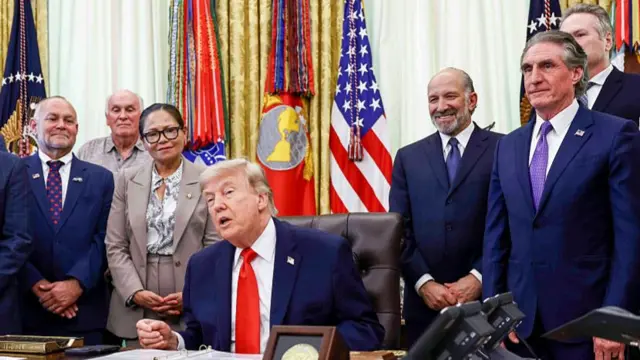 Donald Trump sits behind his desk in the Oval Office surrounded by other people