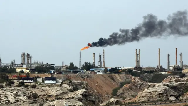 A view of the oil terminal at the Kharg Island. Smoke and flames rising from a large metal structure surrounded by quarries.