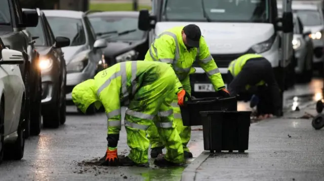 Two workers in yellow hi-viz suits and gloves pick up debris from the road, as a line of cars drives past.