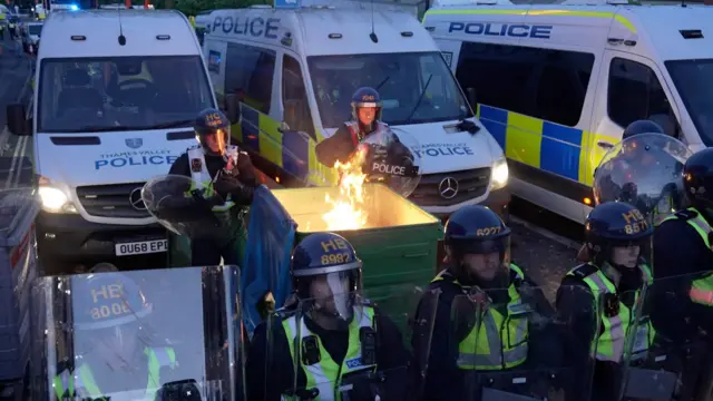 Flaming bin behind a row on police officers in riot gear