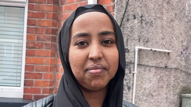 Woman with headscarf, wearing black jacket standing outside house