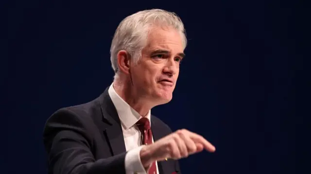 James Cartlidge in a black suit and red tie speaking