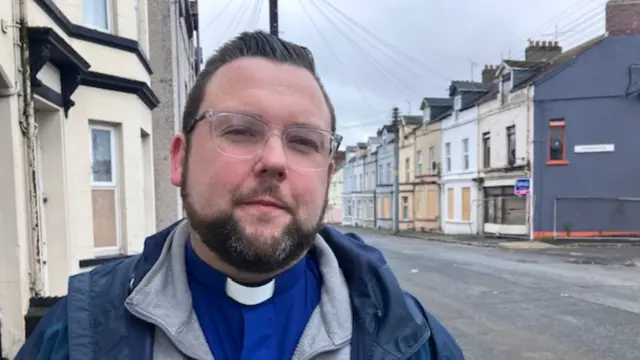 Man with brown hair, glasses wearing a minister collar over a blue shirt. He is wearing a grey fleece and a blue raincoat. He is standing in a street of houses
