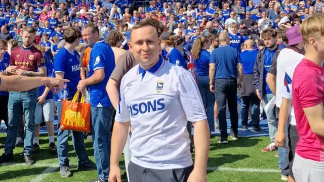 A man wearing a white football shirt stands on a football pitch inside a stadium. Hundreds of other fans mingle around him on the pitch wearing blue football shirts.