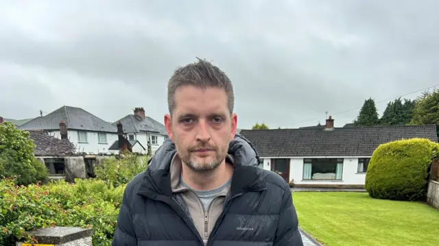 Philip Brett grey hair black coat standing in front of house