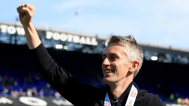 A man with short grey hair on a football pitch within a stadium who is celebrating with one arm raised in the air.