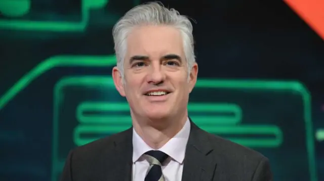 Headshot of James who sits and smiles, with a green and black background behind him. He has white hair and wears a grey suit, pink shirt and gold and black tie
