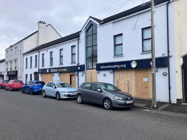 A white building in the middle of a town centre. All pf the windows and doors are boarded up.
