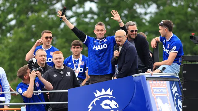 A football team celebrate on an open top bus during a Premier League promotion parade. One man in the middle raises his arms in the air in celebration.