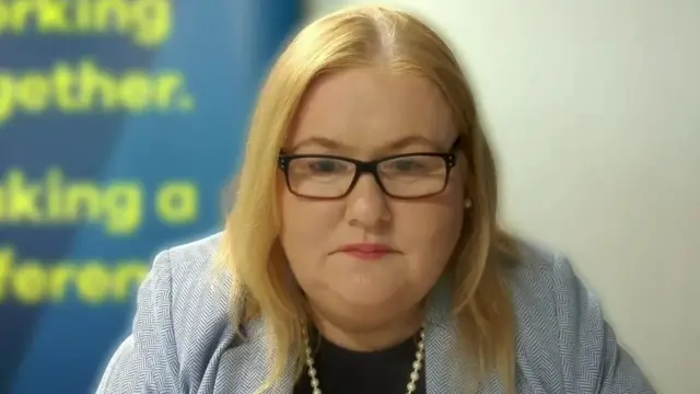 Tracey Reid, a woman with shoulder-length blonde hair, looks towards the camera during a video call. She is wearing a grey suit jacket, a black top and a pearl necklace.