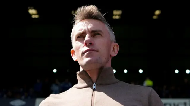 A man stands in the stands of a football stadium looking over the top of the camera toward the pitch. He has short grey hair and is wearing a beige jumper.