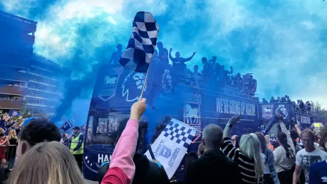 A football club promotion party bus makes it way past crowds who are celebrating. Blue flare smoke billows in the air as fans wave flags.