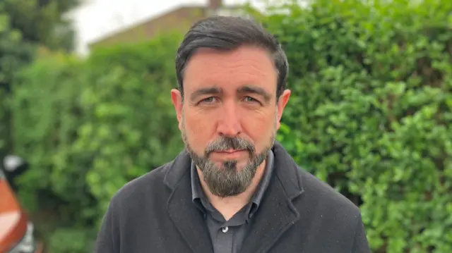 Paul Doherty has dark hair swept over to the side. He has a dark bead which is shot through with grey and is looking at the camera.
