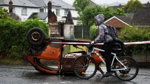 A person dressed in a jacket and black pants rides a white bicycle past an upturned burnt-out orange car that is cordoned off with police tape. The weather is grey and it appears to be raining.