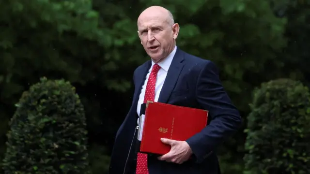 John Healey in a navy suit carrying a small red binder.