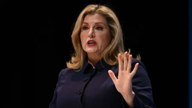 Mourdant speaking in front of a black background with her hand up and palm open