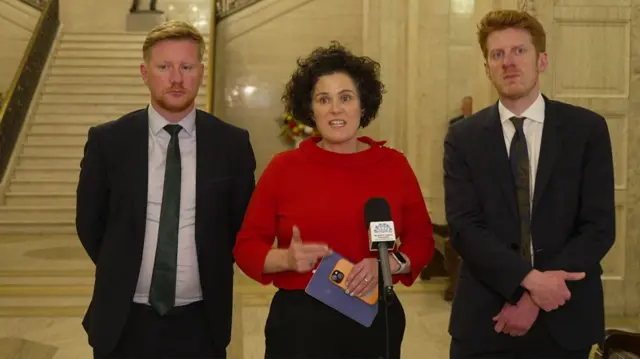 Claire Hanna standing in front of a microphone in Great Hall of Stormont in red top. Daniel McCrossan is to her left wearing a dark suit and green tie. Matthew O'Toole is standing to her right in a dark suit and green tie