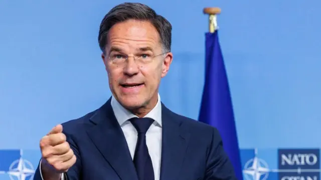 Mark Rutte stands at a podium in a navy suit in front of a blue wall with the Nato flag