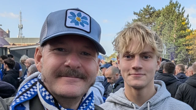 selfie style image of Craig Finbow with his son, Oscar. Craig is wearing a hat and a blue and white scarf. Oscar is wearing a grey hooded top. They are standing in front of a crowd of football fans while looking directly into the camera.