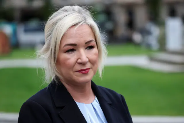 Michelle O'Neill, a woman with straight blonde hair, stands side on to the camera in front of a lawned area. She is wearing a dark suit jacket and a pale blue top.