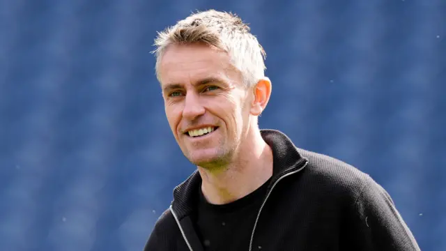 A man with short grey hair stands in a football stadium on a sunny day. He is wearing a black three-quarter length zip jumper with a black T-shirt underneath.