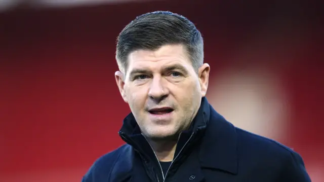 A man with short dark hair wearing a navy coat inside a stadium. He is looking away from the camera and has his mouth slightly open.