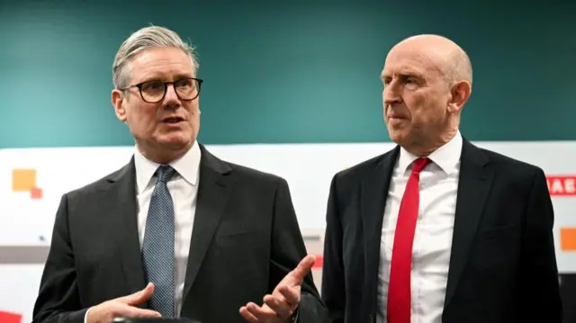 Keir Starmer and John Healey stand side by side wearing black suits