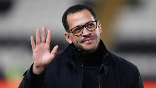 A man with short dark hair wearing glasses and a black coat waves to crowds within a football stadium.