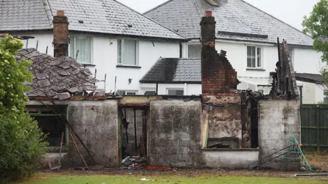 A burnt out building can be seen near Antrim Road. There are no windows and no door and the roof of the building has collapsed. Two chimneys with scorched bricks remain standing