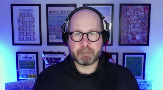 A man wearing headphones, glasses and a black hoodie looks directly into the camera. Posters have been framed on the wall behind him. The room he is in is lit with blue lights.