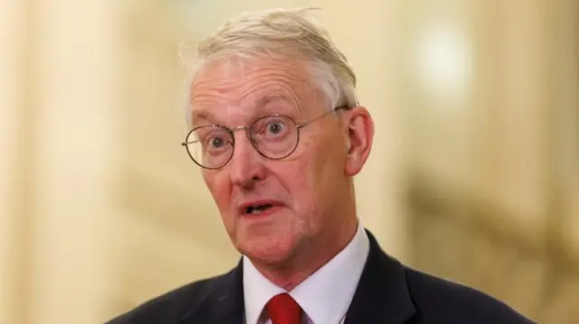 Hilary Benn in a black suit and red tie with round glasses