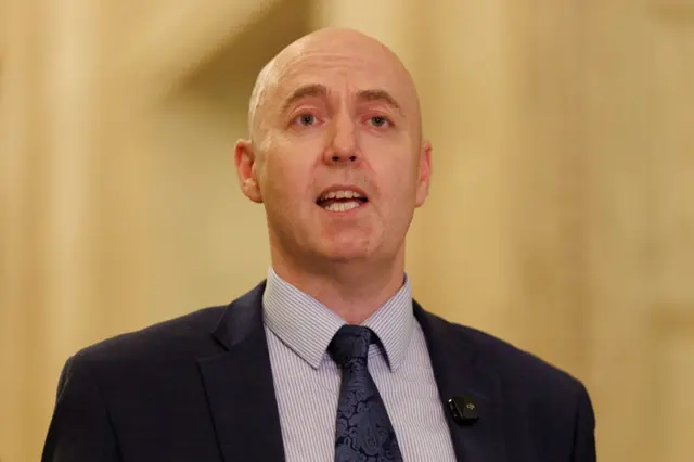 Jon Burrows is speaking in the Great Hall at Stormont. He has a shaved head and is wearing a dark suit with a dark paisley tie