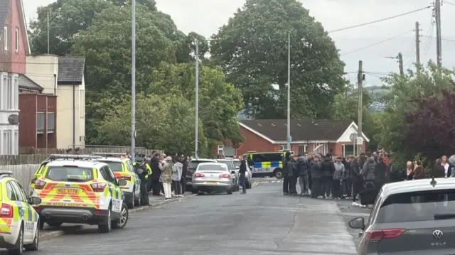 Street- road in centre- psni cars on left side of pavement, grey golf on right side. A small crowd dressed mainly in black are standing on each side of the pavement