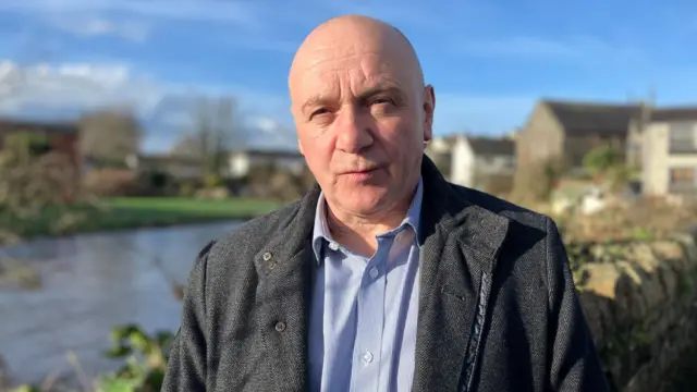 John Blair, wearing a blue shirt and dark grey jacket. He is standing in front of a river with a blue sky above.
