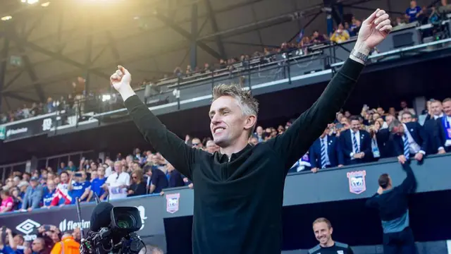 Ipswich Town manager Kieran McKenna smiling and celebrating with both arms raised in victory, wearing a black quarter-zip jumper in front of cheering fans at a packed football stadium.
