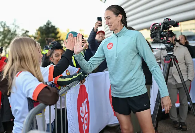 Jill Scott is wearing black shorts and a light blue top with a quarter zip and the Sport Relief logo which is a red circle with white writing. Her brown hair is in a plait down her back. She is high-fiving some children who are wearing white and red tops. There is a camera on a tripod behind her