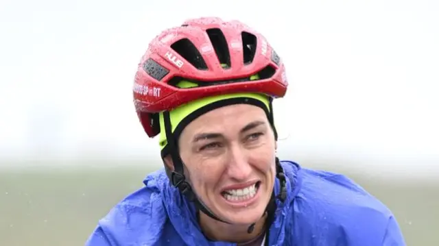 Close up of Jill Scott cycling. She is wearing a red helmet and a blue raincoat. Raindrops are falling off her. She is bearing her teeth in exertion.