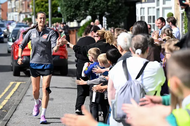 Jill Scott is running on a pavement and is high-fiving with a crowd of people standing on the pavement. It is sunny.