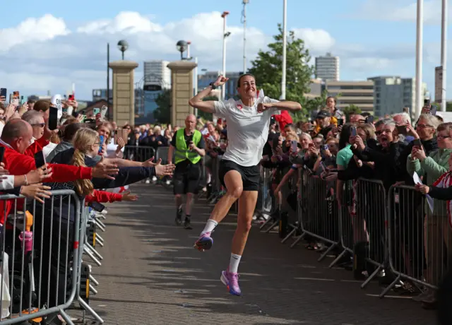 Jill Scott jumps in the air as she crosses the finish line at the Stadium of Light. She is wearing black shorts and a white T-shirt. Crowds of people are lining both side of the finish funnel and cheering