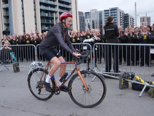 Jill Scott sets off from Wembley Stadium. She is on a bike with a brown frame and black tyres. She is wearing a red cycle helmet, black shorts and a black top. There are metal barriers and groups of children behind her.