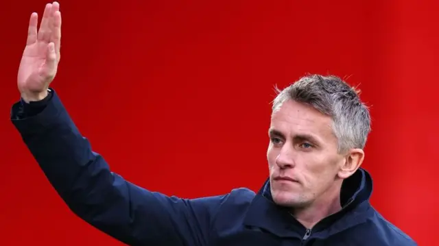 Kieran McKenna, with short grey hair, stands against a bright red background, looking to the side with a serious expression. He is wearing a dark navy blue buttoned jacket with a small circular badge on the left side of his chest, and his right hand is raised with his palm facing forward as if waving or gesturing.