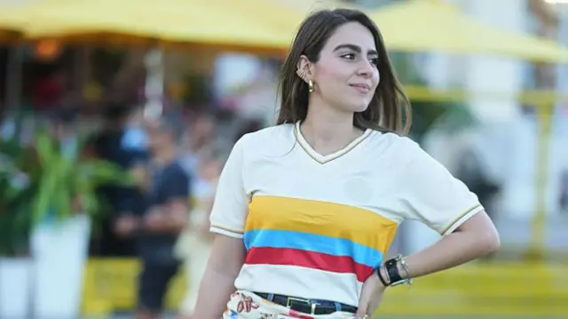 A guest is seen wearing gold earrings, a white, yellow, red, and blue short-sleeved Colombia national team jersey, a black belt, a cream and multicolored patterned scarf wrapped around the waist, blue denim shorts, and white sneakers at the FIFA World Cup 2026 Kick-Off Watch Party on June 11, 2026 in Miami Beach, Florida