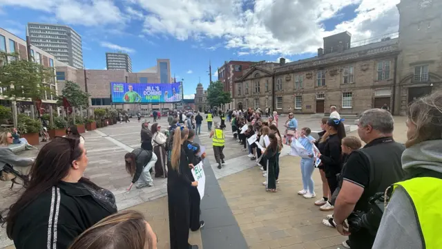 Rows of people lining up in Keel Square in Sunderland. There are two rows facing each other with a space in the middle. They are holding posters. In the background is an electronic billboard with a big picture of Jill Scott and the message Jill's coming home